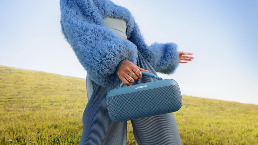 A person in a meadow holding a blue Bose SoundLink Max speaker. 