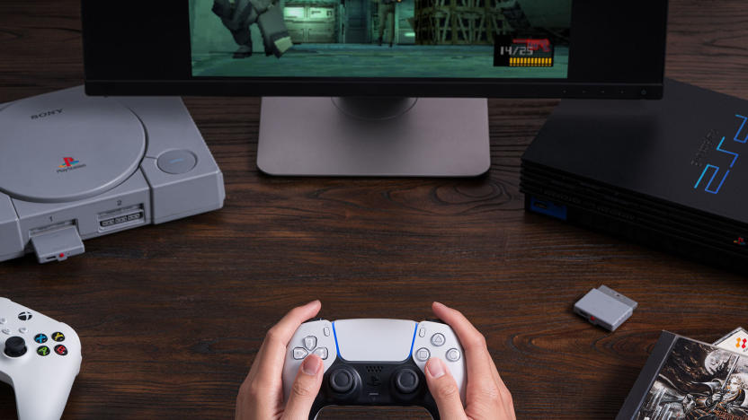 A pair of hands holds a PlayStation 5 DualSense controller. A PlayStation 1 and PlayStation 2 are in the background and a dongle sits on the table. A game is shown on a monitor.