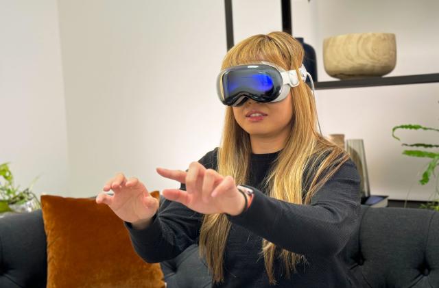 A woman with blonde hair with the Apple Vision Pro on her face, her hands are outstretched with her fingers held in a typing position.