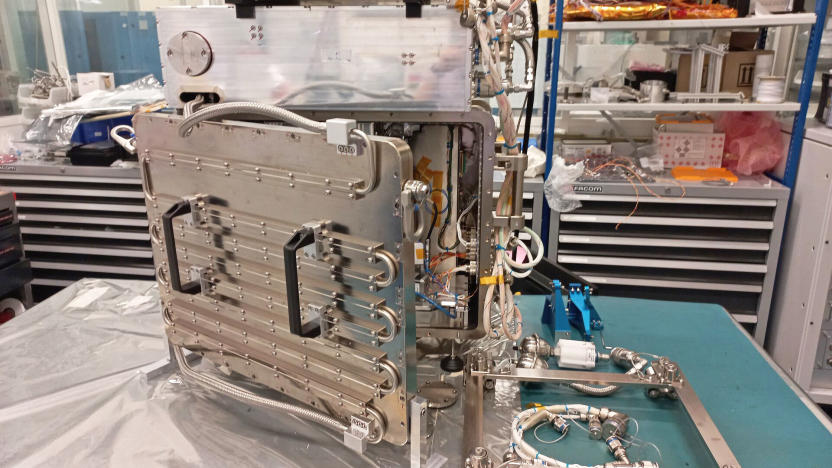 A raw metal 3D printer with lots of stainless steel and tubing that will be tested on the International Space Station sits on a cluttered workbench in a lab.