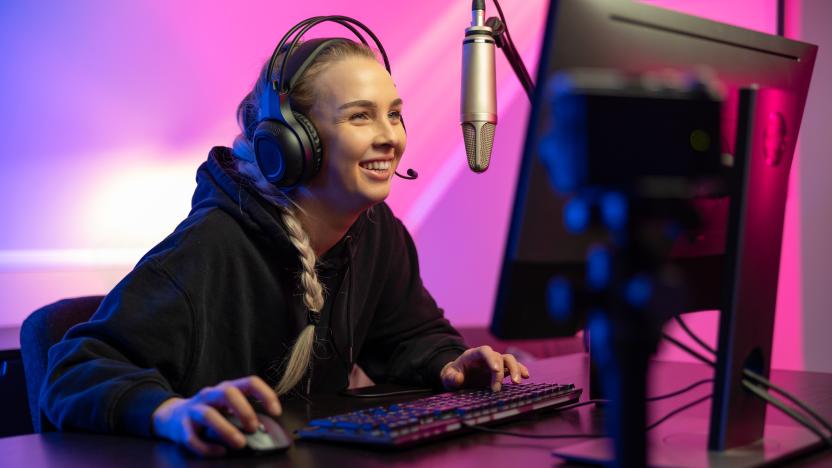 Smiling and excited professional blonde esport gamer woman with headphones streaming vlog live while playing online video game on her PC. Room with blue and pink colored LED lights.
