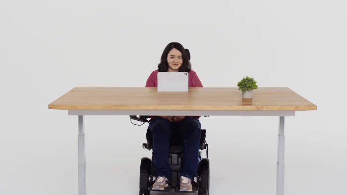 A person in a wheelchair sitting in front of a table where an iPad is set up. They are facing the iPad and smiling.