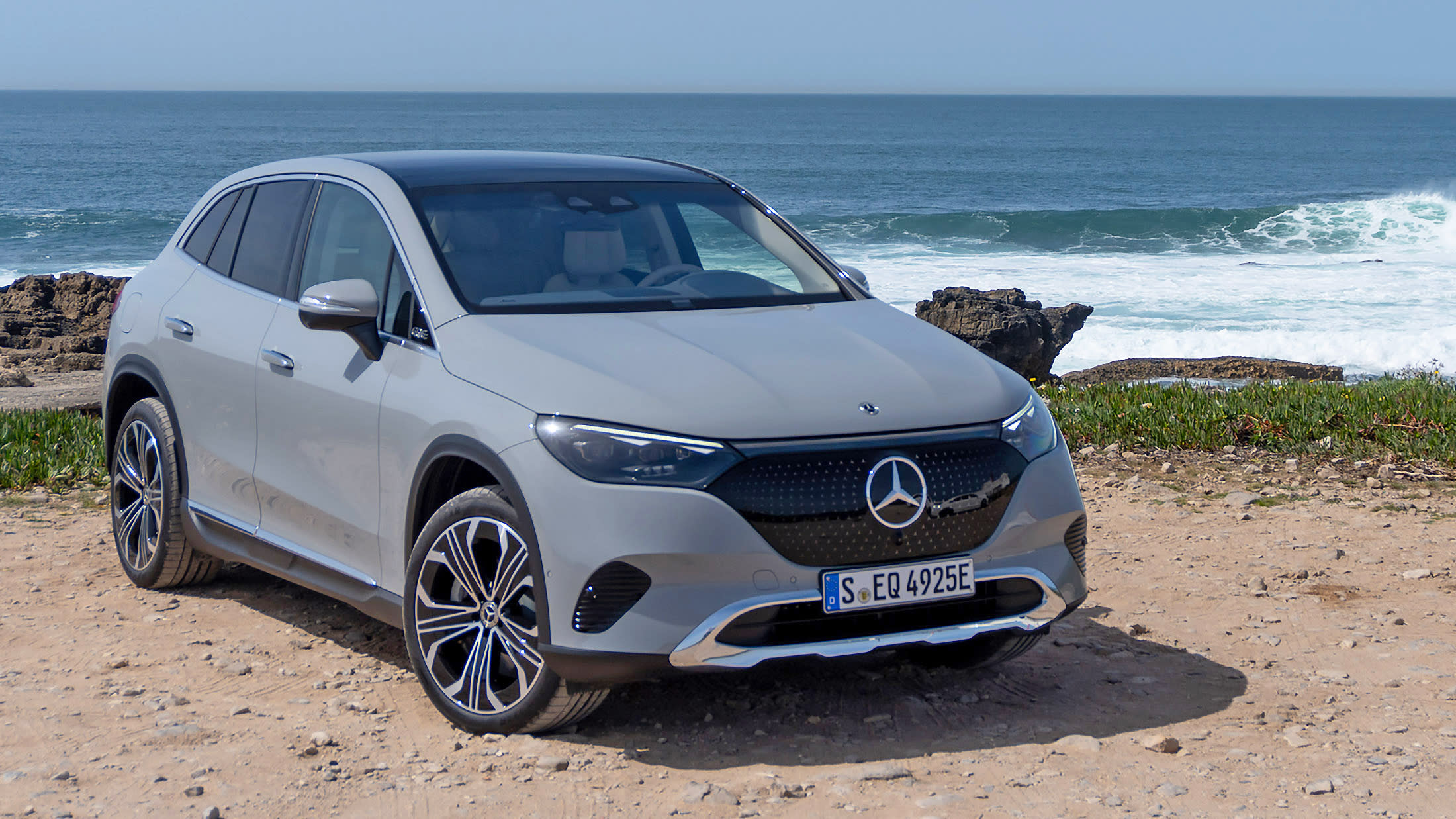 A grey Mercedes EQE SUV is parked on a rocky shore with rock outcroppings and waves close at hand in the background.