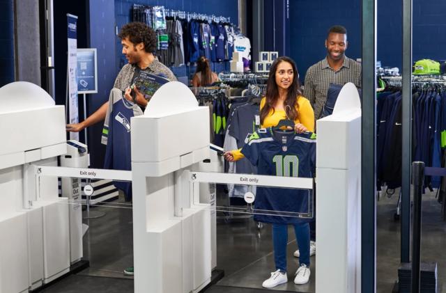 Customers use Amazon’s Just Walk Out tech in a merch store at Seattle’s Lumen Field. Three customers walk smiling as they prepare to exit through turnstiles.