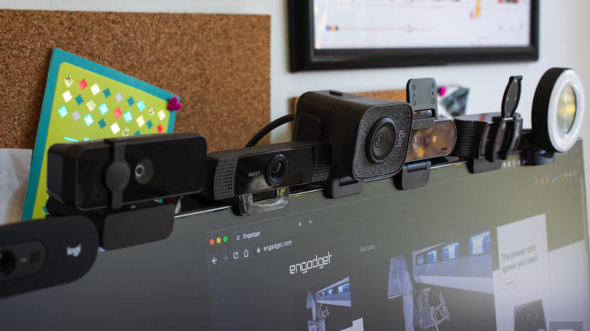 Seven webcams sitting on top of a computer monitor with cork boards on the wall behind them.