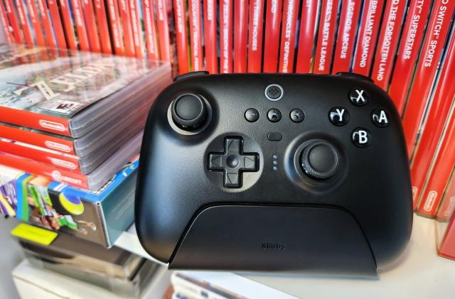 A black video game controller, the 8BitDo Ultimate Bluetooth Controller, rests on a white shelf in front of a row of several red Nintendo Switch video game cases.