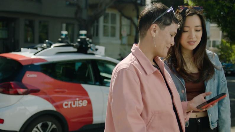 Two persons in the foreground, both looking down on a smartphone. In the background is a Cruise robotaxi painted red and white.