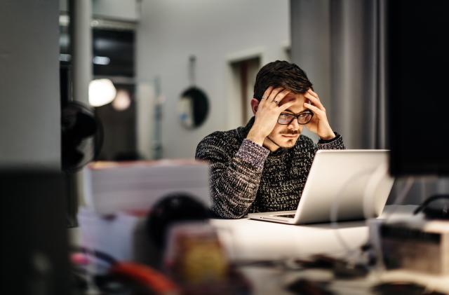 Young casual businessman wearing glasses is sitting in front of his notebook holding his head pondering over his work. Office equipment and another computer is in front of him.