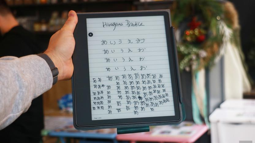 The Amazon Kindle Scribe held up in mid-air by a hand gripping its right side. The screen shows the words "Hiragana Practice" at the top, followed by columns of Japanese script below.