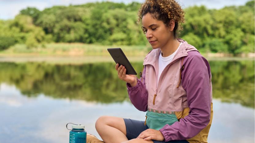 A woman sits on a rock near a lake reading from an Amazon Kindle