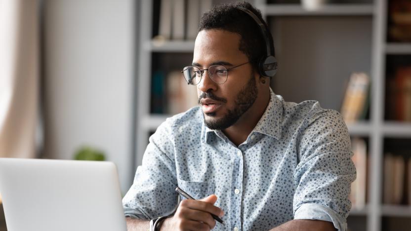 Focused young african businessman wear headphones study online watching webinar podcast on laptop listening learning education course conference calling make notes sit at work desk, elearning concept