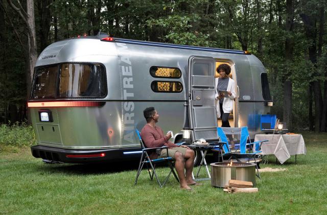 The Airstream eStream electric concept camper parked on the grass by a tree line with a couple sitting outside of it in camping chairs.