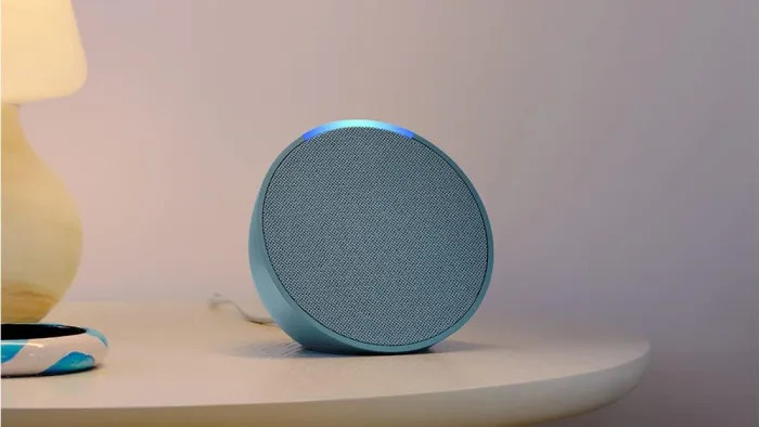 The Amazon Echo Pop speaker in blue sits on a night table with a yellow lamp nearby. 