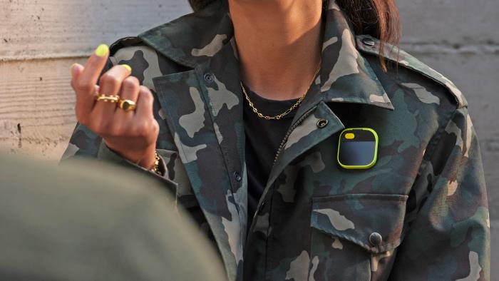 A cropped image showing the torso of a women wearing a camouflage jacket with a yellow Humane AI Pin on the chest.