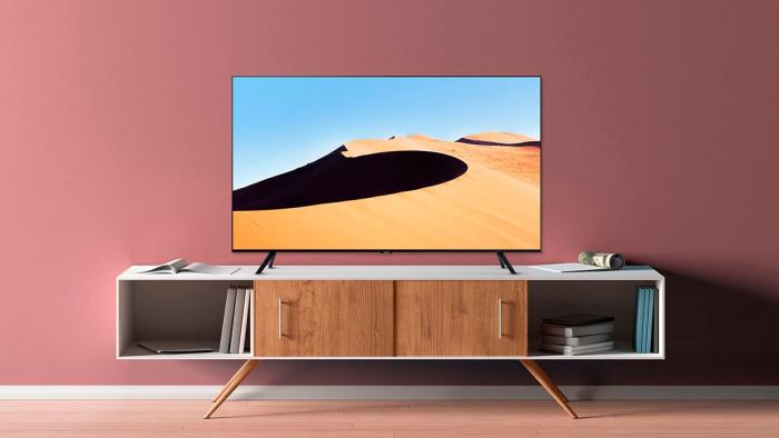 A press image of the Samsung TU690T 4K TV, displaying a TV on a white and brown cabinet against a light red wall. The image on the TV displays a desert dune.