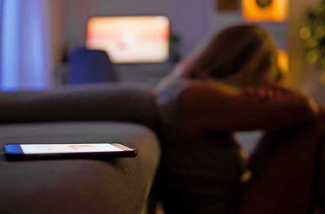 A seemingly depressed woman sits on floor in front of a couch hugging her knews, with a mobile phone in the foreground on the sofa.