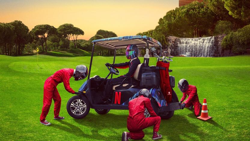 A person wearing a Formula 1 helmet sits in a golf buggy on a golf course as technicians swap the wheels.