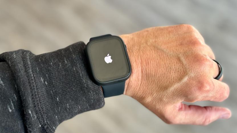 A left arm / wrist / hand wearing an Apple Watch with a white Apple logo on its otherwise black screen. Blurred gray background.