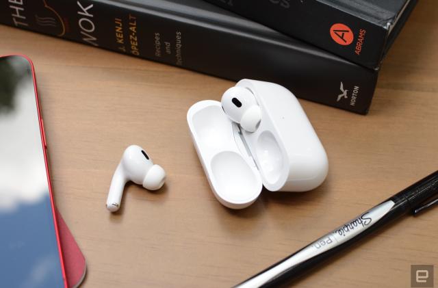 Apple's white AirPods Pro and case sit on a wooden table with books, an iPhone and a pen nearby. One of the earbuds has been removed from the case and sits next to it. 