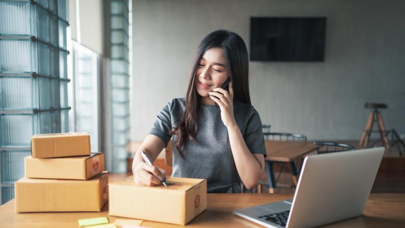 Young business woman working selling online. Entrepreneur owner using smartphone or laptop taking receive and checking online purchase shopping order to preparing pack product box. Packing goods for delivery to customer. Online selling. E-commerce. Online Shopping