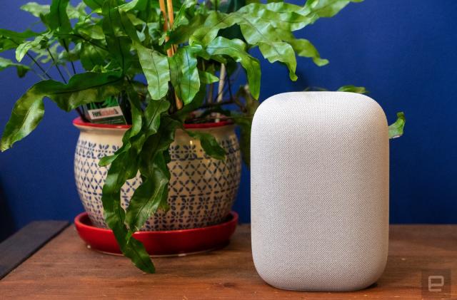 Google Nest Audio smart speaker sitting on a wooden tablet next to a green plant, against a blue wall.