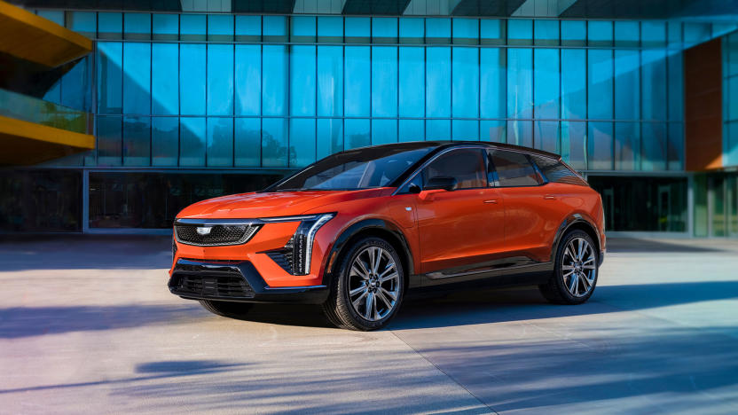 Marketing photo for the Cadillac Optiq. It shows the compact SUV in orange sitting in front of a modern luxury home or garage.