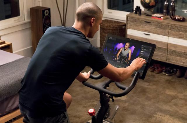 A promotional image of a man in his room riding a Peloton bike with a touchscreen display in front.