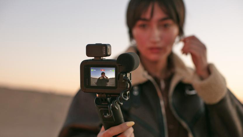 A woman takes a selfie using the GoPro Volta Battery Grip while standing on a beach or with a sand dune behind her.