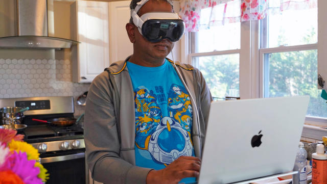 A man stands in front of his laptop in a kitchen while wearing the Apple Vision Pro AR/VR headset.
