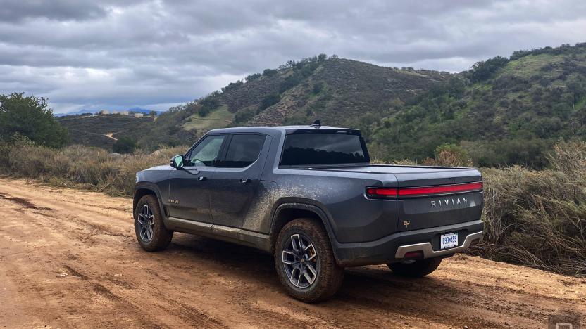 A dark grey Rivian R1T drives up a dirt road that's headed upwards with hills in the background.