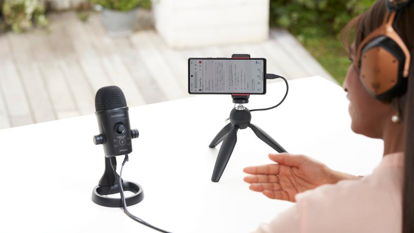 Lifestyle marketing photo of a person (view from behind, only part of them is visible in the right of the frame) talking into a Rode Go:Podcast microphone with a smartphone set up on a mini tripod next to it. White table with a blurred environment behind it.