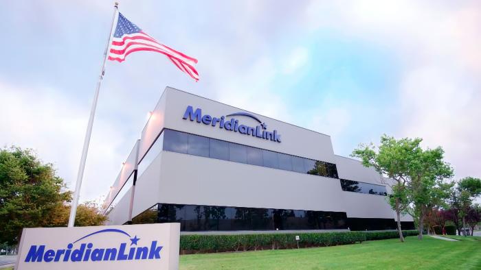 Company photo of MeridianLink headquarters. A modern building with company logo behind a green lawn with a sign (also bearing the company logo) and an American flag.