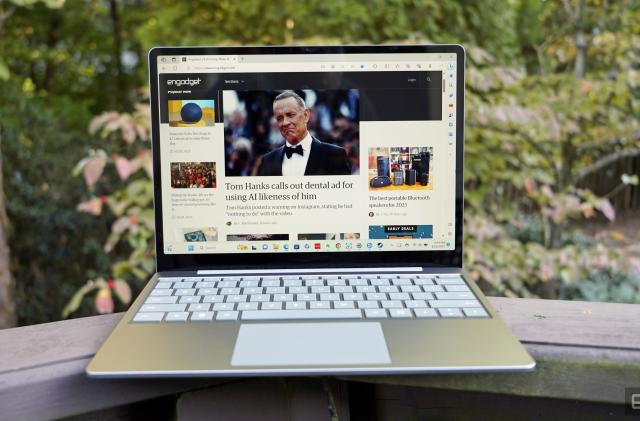 A Surface Laptop Go 3 sits open on a backyard deck railing with foliage in the background.