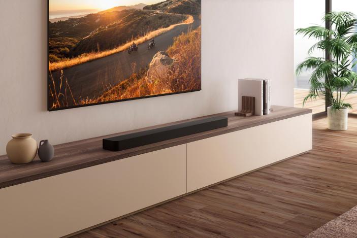 A black soundbar on a wooden shelf underneath a TV with a mountain road sunset scene on the display.