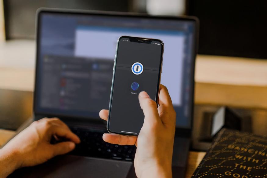 A person holding an iPhone with the 1Password app open. A laptop is in the background.