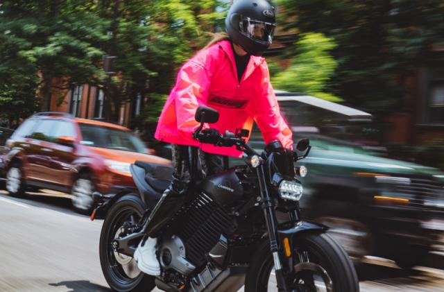 A person in a flamingo-pink windbreaker riding the LiveWire S2 Del Mar electric motorcycle down a city street. Cars are parked behind in front of trees and visible sections of city buildings.