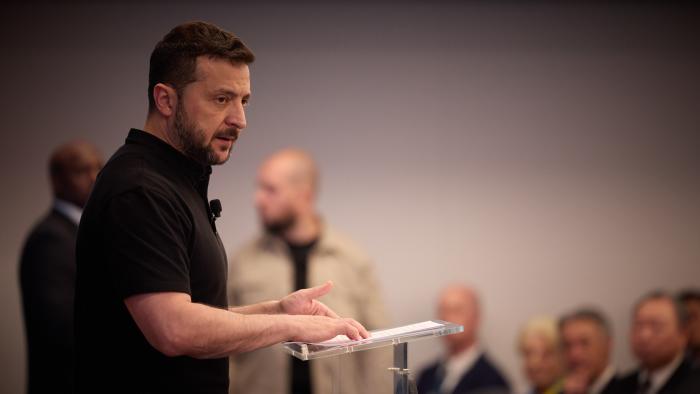 Volodymyr Zelenskyy standing at a podium addressing an audience of government allies.