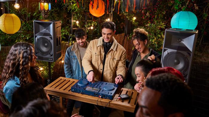 A DJ spins on an Omnis-Duo controller flanked by two Wave-Eight speakers amidst a small but lively party in a backyard.