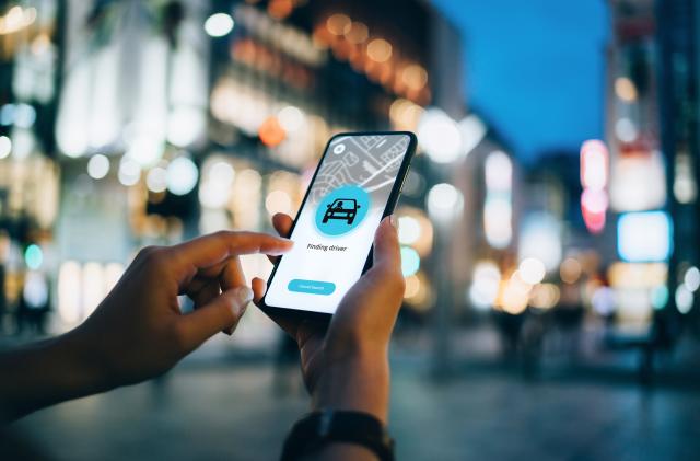 Close up of young woman using mobile app device on smartphone to arrange a taxi ride in downtown city street, with illuminated city traffic scene as background