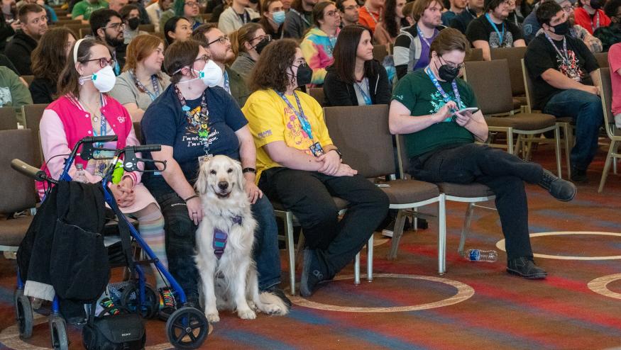 A room full of audience members at a Games Done Quick charity speed-running event. In the front row, someone holds a cream-colored golden retriever, who smiles for the camera.
