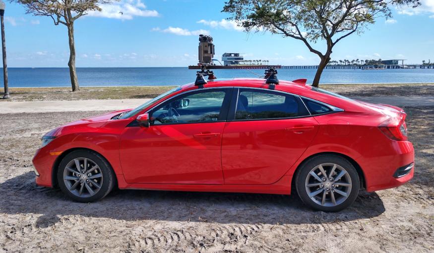 A red car parked on the sand near a waterfront has the new Google Street View camera system mounted on its roof.
