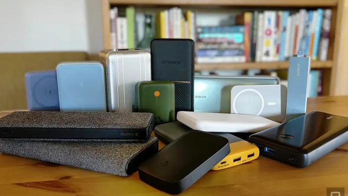 Fourteen different power banks and battery packs from brands like Anker, Otterbox, and Mophie arranged on a wooden table with a bookcase in the background. 