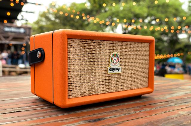 The Orange Box portable Bluetooth speaker sits on a wooden picnic table with string lights overhead, with a crowded outdoor event space and trees blurred in the background.