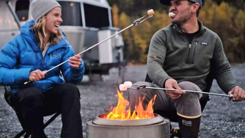 A Solo Stove firepit burns in an outdoor setting as two people toast marshmallows in its flames. A camper trailer is in the background.  