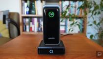 The Anker Prime power bank sits on its charging dock atop a wooden table. There are books and plants in the background. 