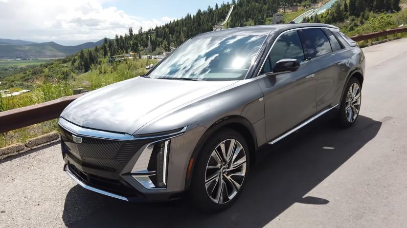 A gray Cadillac Lyriq driving on a hillside road with green trees in the background. Photo taken from the front left of the vehicle.