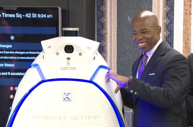 NYC mayor Eric Adams makes one half of a heart sign with his hand against a robot that has no arms.