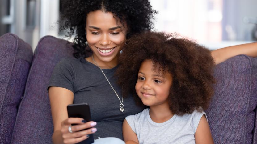 Happy afro mother or elder sister with little kid sitting on sofa in living room at home spend time using phone app having fun, watching cartoons make videocall, modern tech weekend activities concept