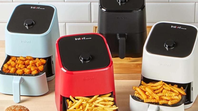 A collection of compact air fryers in various colors sit on a wooden countertop against a white tiled wall, with their bottom compartments open, revealed cooked snacks like french fries and tater tots.