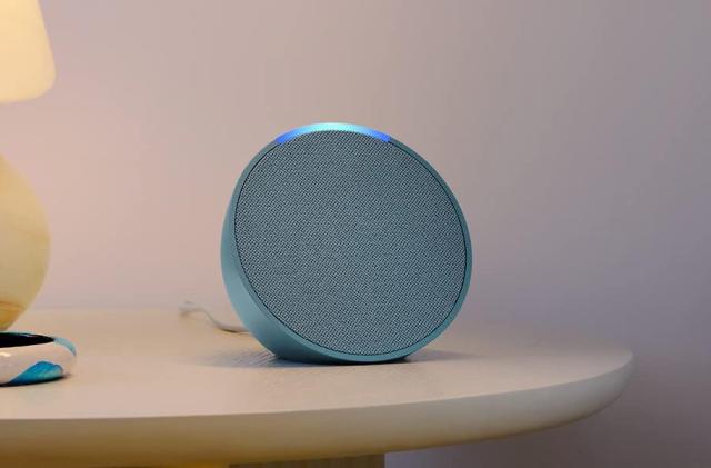 The Amazon Echo Pop speaker in blue sits on a night table with a yellow lamp nearby. 